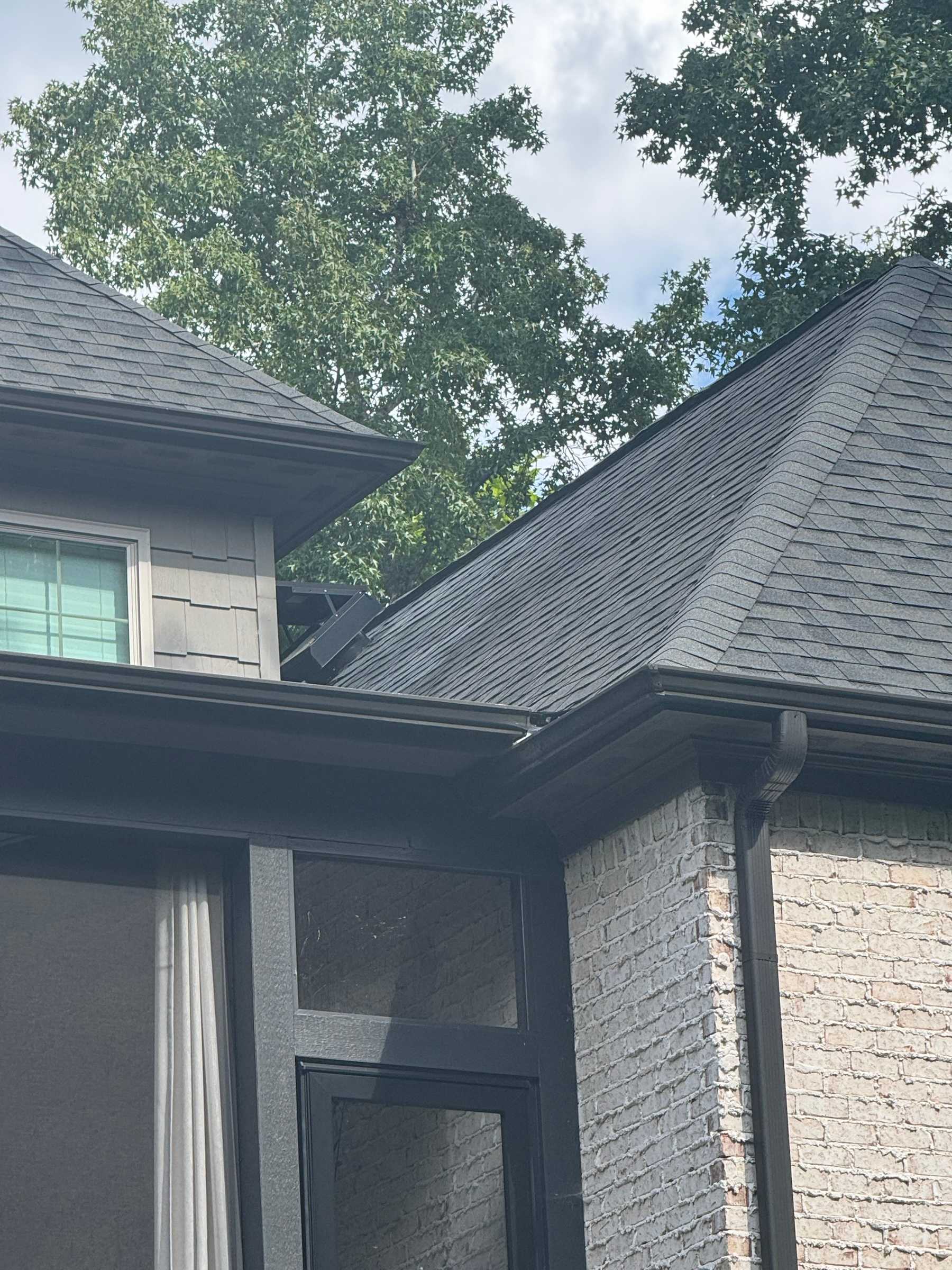 Close-up of new black gutters and a downspout under a dark shingle roof on a painted brick house.