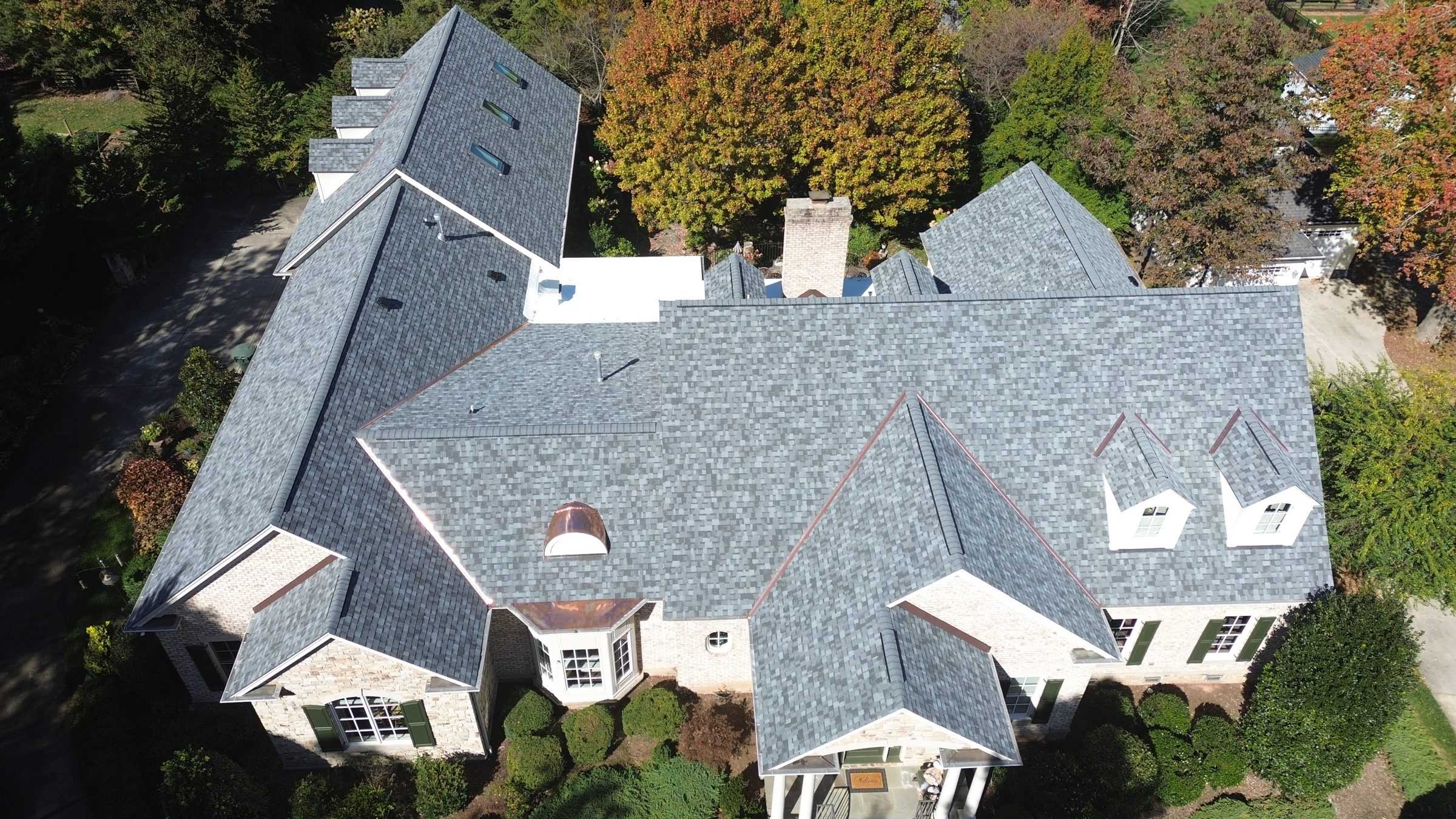 Drone view of a large house among trees with a new gray roof and many valleys.