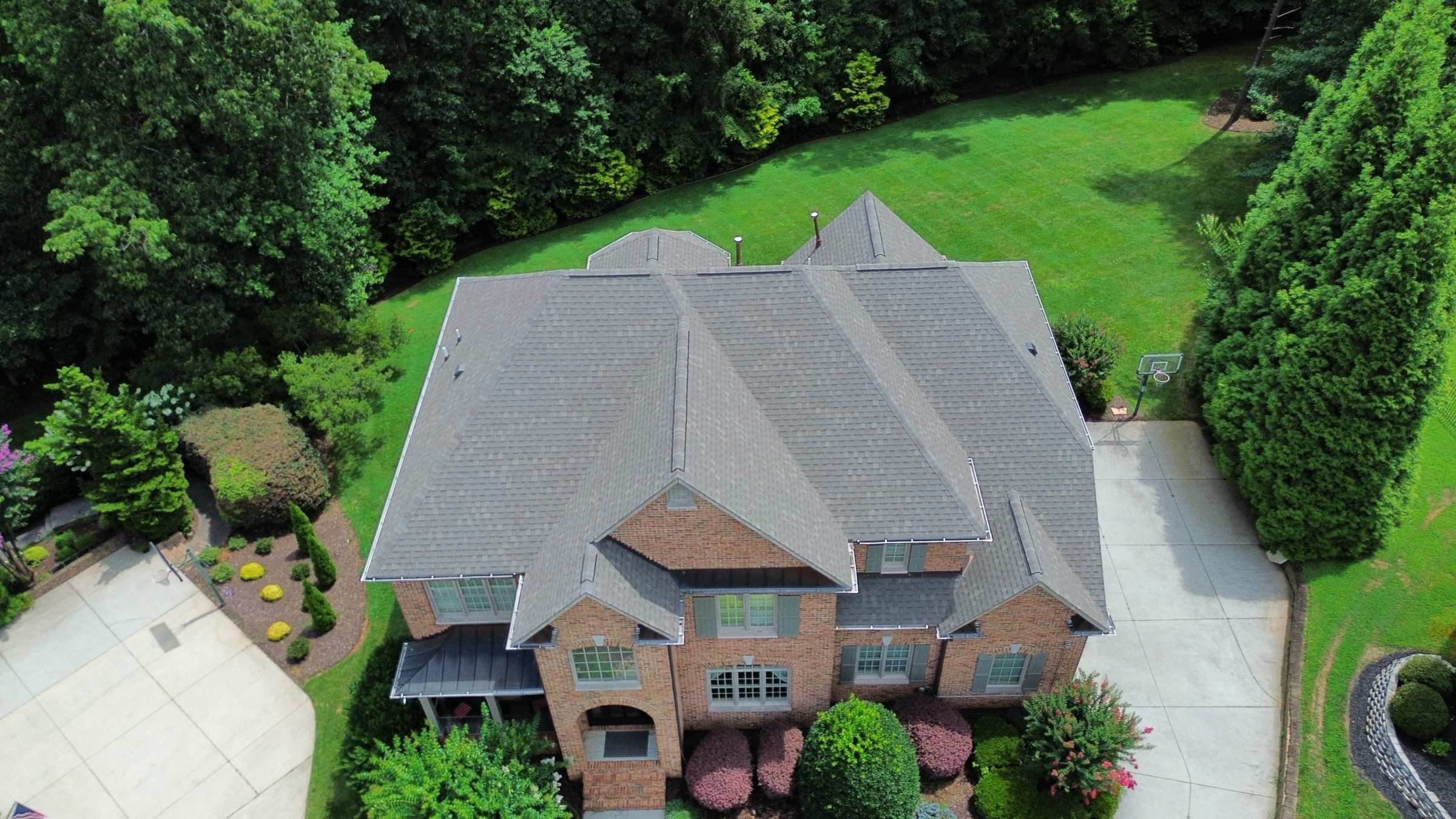 Drone view of a large brick house with a new gray shingle roof and a wide lawn behind it.