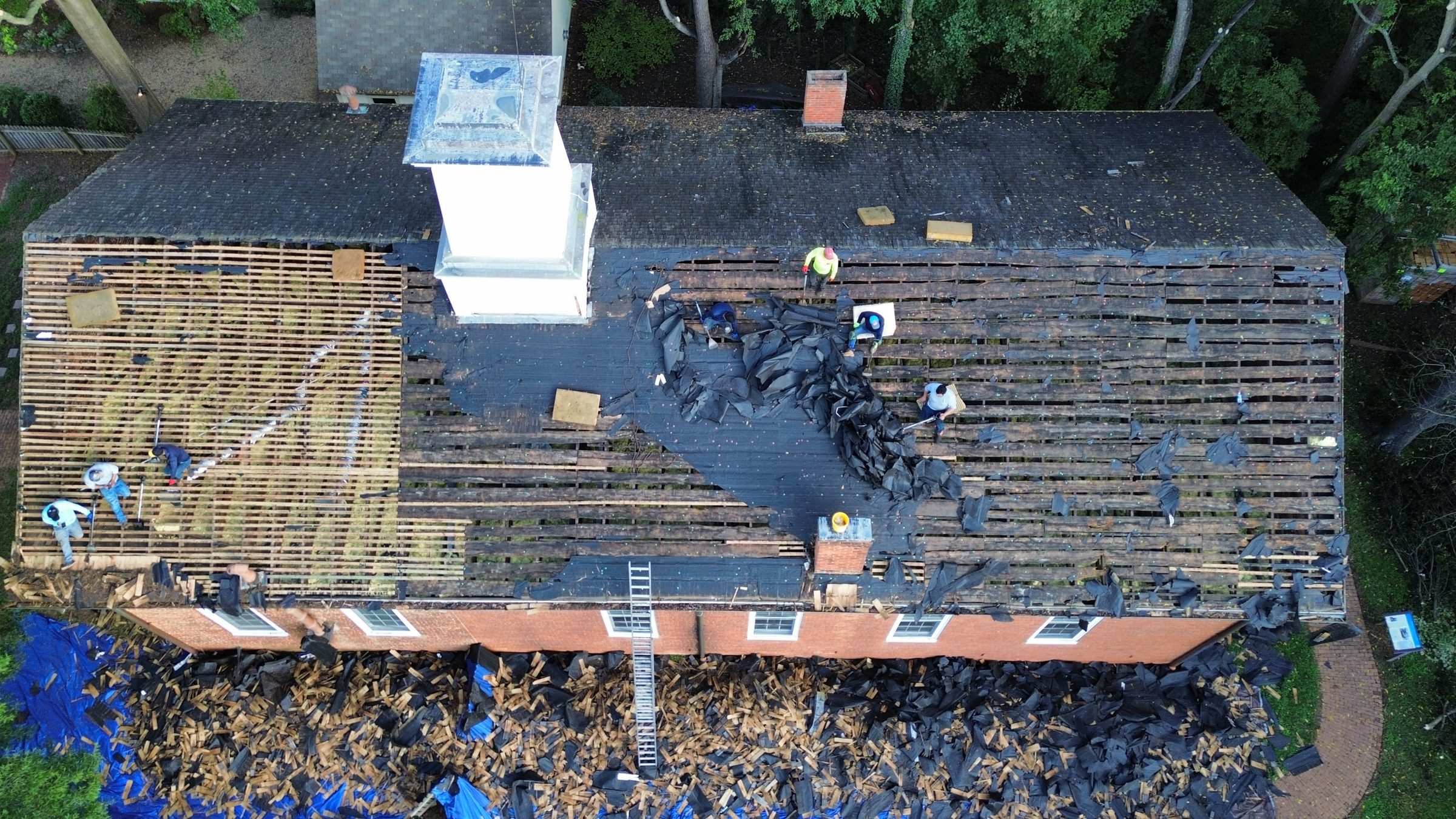 Close drone view of a crew stripping a roof down to the boards, with tarps below.
